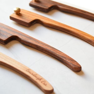 May include: Four wooden handled knives with brass accents. The handles are made of different types of wood, including a dark wood with a striped pattern. The knives are arranged in a row, with the handles pointing to the right. The text "Sawdaugh & Co." is engraved on the handle of one of the knives.
