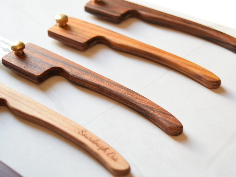 May include: Four wooden handled knives with brass accents. The handles are made of different types of wood, including a dark wood with a striped pattern. The knives are arranged in a row, with the handles pointing to the right. The text "Sawdaugh & Co." is engraved on the handle of one of the knives.