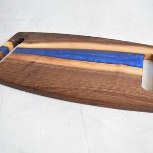 Op de afbeelding: Een lang, rechthoekig serveerplank gemaakt van walnoot hout met een blauwe epoxyhars inlay. De plank heeft twee uitgesneden handvatten aan beide zijden.