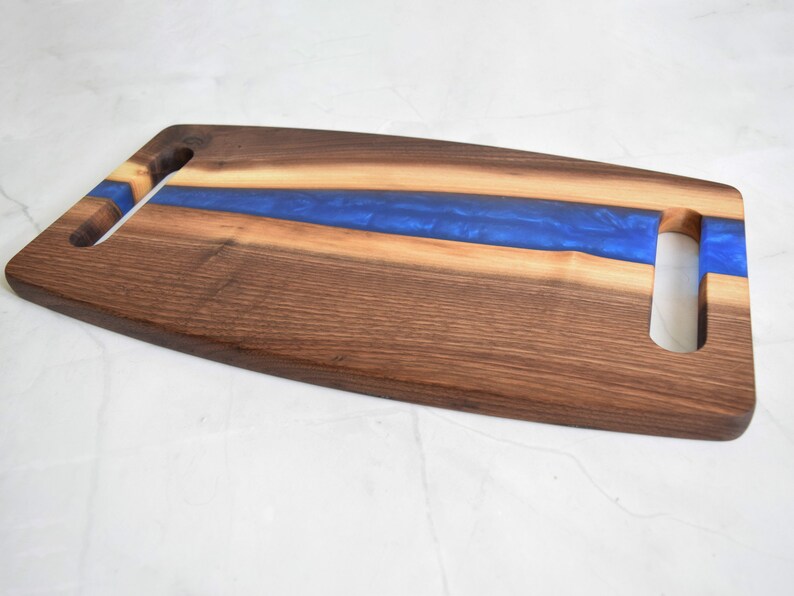 Op de afbeelding: Een lang, rechthoekig serveerplank gemaakt van walnoot hout met een blauwe epoxyhars inlay. De plank heeft twee uitgesneden handvatten aan beide zijden.