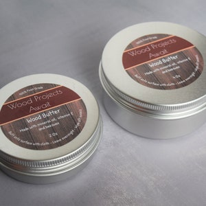 May include: Two silver metal tins with labels that read "Wood Projects Await" and "Wood Butter". The labels are brown with white text and feature a wood grain pattern. The smaller tin is labelled "2 oz" and the larger tin is labelled "4 oz".