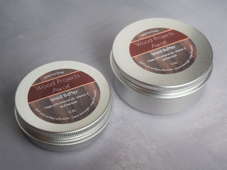 May include: Two silver metal tins with labels that read "Wood Projects Await" and "Wood Butter". The labels are brown with white text and feature a wood grain pattern. The smaller tin is labelled "2 oz" and the larger tin is labelled "4 oz".