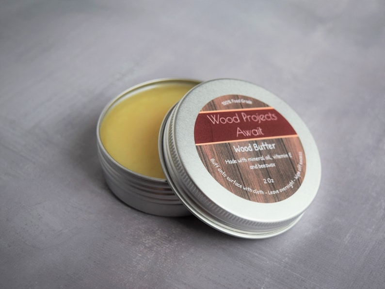May include: A small silver tin with a label that reads "Wood Projects Await Wood Butter Made with mineral oil, vitamin E and beeswax 2 oz Buff onto surface with cloth - Leave overnight". The tin is open and contains a yellow, waxy substance.