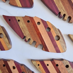 May include: A selection of handcrafted wooden cheese knives. Each knife has a unique shape and is made from alternating strips of reddish-purple, light brown, and medium brown wood. The knives have circular and slotted cutouts.