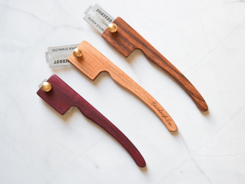 May include: Three wooden straight razors with a metal blade and a brass knob. The razors are made of different types of wood, including maple, walnut, and purpleheart. The razors are engraved with the words "Sourdough Era" and "Shaverb".