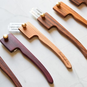 May include: Four wooden bread lame tools with different colored handles. The handles are made of dark wood, light wood, purple wood, and a striped wood. Each lame has a razor blade attached to the top. The blades are labeled "Shavet" and "Derby". The light wood lame has the words "Sourdough Era" engraved on the handle.
