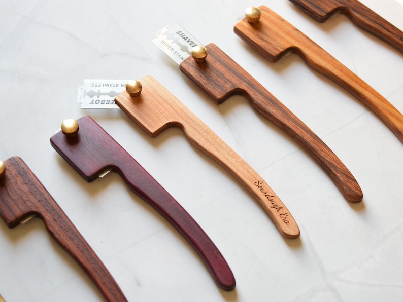 May include: Four wooden bread lame tools with different colored handles. The handles are made of dark wood, light wood, purple wood, and a striped wood. Each lame has a razor blade attached to the top. The blades are labeled "Shavet" and "Derby". The light wood lame has the words "Sourdough Era" engraved on the handle.