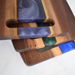 Op de afbeelding: Een stapel van drie houten snijplanken met verschillende gekleurde epoxyharsinleg. De bovenste plank heeft een paarse inleg, de middelste plank heeft een groene inleg en de onderste plank heeft een blauwe inleg.