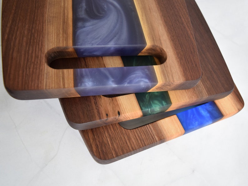 Op de afbeelding: Een stapel van drie houten snijplanken met verschillende gekleurde epoxyharsinleg. De bovenste plank heeft een paarse inleg, de middelste plank heeft een groene inleg en de onderste plank heeft een blauwe inleg.