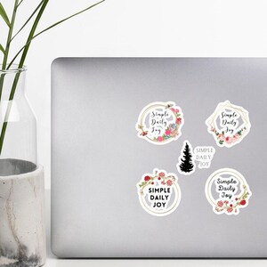 Può includere: Cinque adesivi con un design floreale e il testo "Simple Daily Joy" in un cerchio. Gli adesivi sono su un laptop argentato.