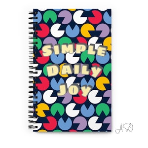 Puede incluir: Una libreta de espiral con una cubierta azul que presenta un patrón geométrico colorido de círculos y formas de Pac-Man. El texto "SIMPLE DAILY JOY" está impreso en letras amarillas y grises en la cubierta.
