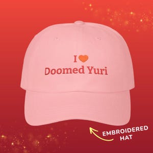 Könnte beinhalten: Eine hellrosa Baseballkappe mit dem gestickten roten Text "I", gefolgt von einem orangefarbenen Herzsymbol und dem roten Text "Doomed Yuri", mittig auf der Vorderseite. Die Kappe hat einen gebogenen Schirm und eine strukturierte Krone.