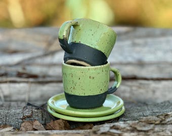 May include: Two green ceramic mugs with black bands stacked on top of each other. The mugs are sitting on two green ceramic saucers.