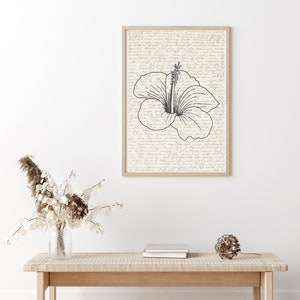 Puede incluir: Una impresi&oacute;n art&iacute;stica enmarcada con una ilustraci&oacute;n de flor de hibisco en blanco y negro. La obra de arte est&aacute; sobre un fondo de escritura descolorida, encerrada en un marco de madera clara. La impresi&oacute;n se muestra sobre un banco de madera clara con una parte superior tejida.