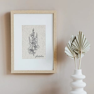 May include: A framed print of a black and white line drawing of a gladiolus flower on a vintage background. The frame is light wood and the print is hanging on a light beige wall. A white vase with dried palm leaves is on a white marble surface.