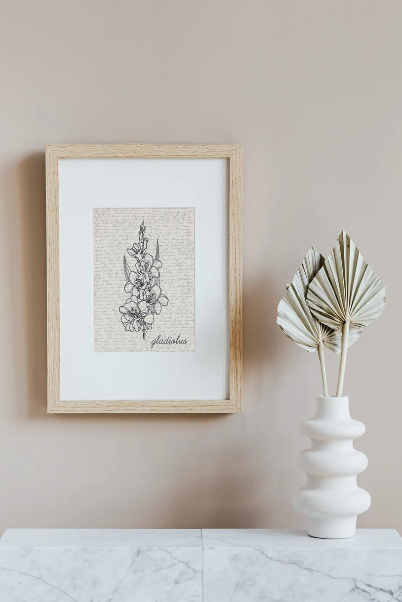 May include: A framed print of a black and white line drawing of a gladiolus flower on a vintage background. The frame is light wood and the print is hanging on a light beige wall. A white vase with dried palm leaves is on a white marble surface.