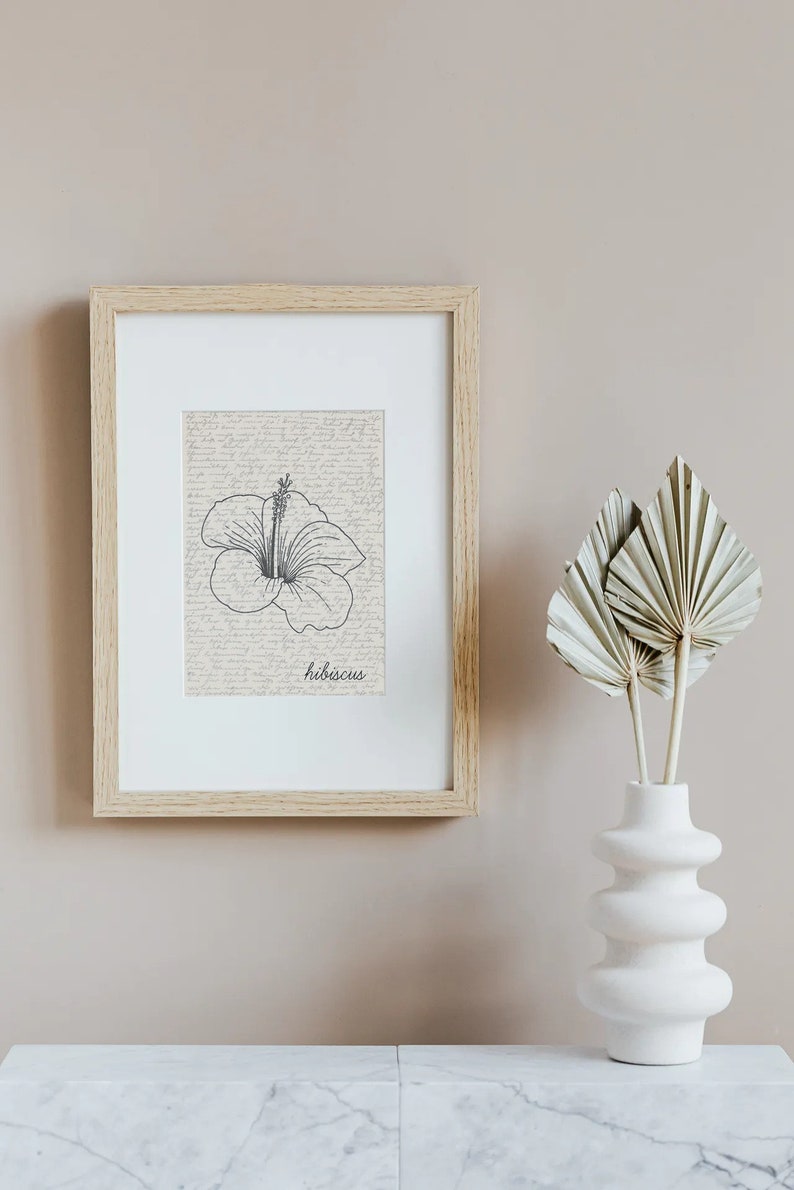 Puede incluir: Una impresi&oacute;n bot&aacute;nica enmarcada con una ilustraci&oacute;n de flor de hibisco en blanco y negro y la palabra "hibiscus". La impresi&oacute;n est&aacute; en un marco de madera clara. Un jarr&oacute;n blanco con hojas de palma secas est&aacute; al lado del marco.