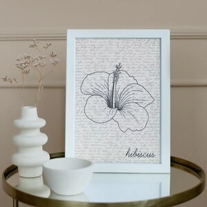 Puede incluir: Una impresi&oacute;n enmarcada en blanco con una ilustraci&oacute;n de flor de hibisco en blanco y negro con la palabra "hibiscus" en escritura. El fondo tiene un dise&ntilde;o de escritura vintage. Un jarr&oacute;n y un cuenco blancos se encuentran sobre una mesa dorada.