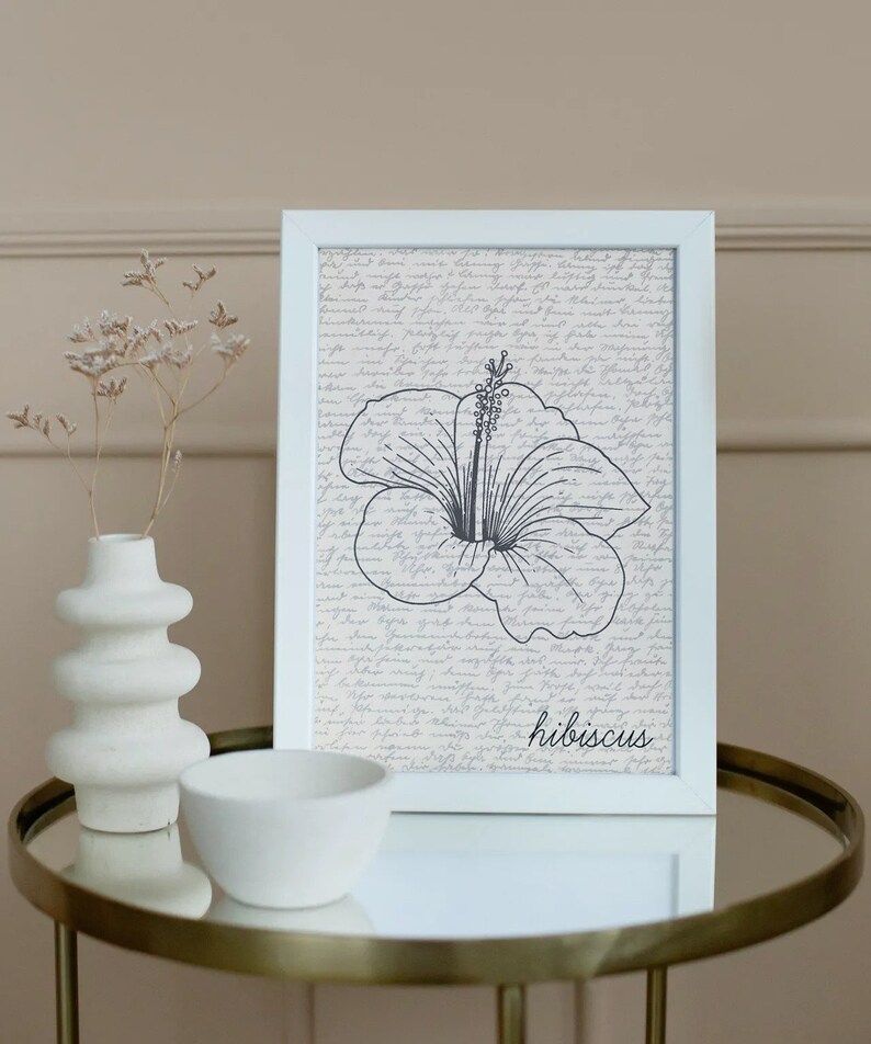 Puede incluir: Una impresi&oacute;n enmarcada en blanco con una ilustraci&oacute;n de flor de hibisco en blanco y negro con la palabra "hibiscus" en escritura. El fondo tiene un dise&ntilde;o de escritura vintage. Un jarr&oacute;n y un cuenco blancos se encuentran sobre una mesa dorada.