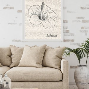 Puede incluir: Una impresi&oacute;n art&iacute;stica enmarcada con una ilustraci&oacute;n en blanco y negro de una flor de hibisco. La obra de arte est&aacute; sobre un fondo de escritura a mano. La palabra "hibiscus" est&aacute; escrita en cursiva. La impresi&oacute;n se muestra sobre un sof&aacute; beige.