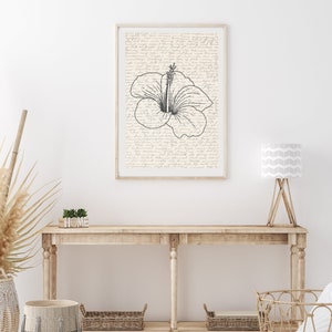 Puede incluir: Una impresi&oacute;n art&iacute;stica enmarcada con una ilustraci&oacute;n de flor de hibisco en blanco y negro. El fondo es de color crema con escritura manuscrita. La obra de arte se exhibe sobre una consola de madera con cestas y una l&aacute;mpara.