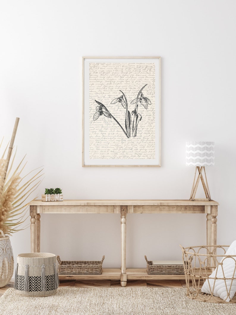 K&ouml;nnte beinhalten: Eine Schwarzwei&szlig;illustration von Schneegl&ouml;ckchen, die auf einem cremefarbenen Hintergrund mit handschriftlichem Text gedruckt ist. Der Druck ist gerahmt und h&auml;ngt an einer wei&szlig;en Wand &uuml;ber einem Holz-Konsolentisch.