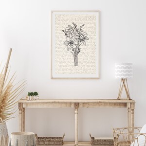May include: A black and white line drawing of a bouquet of flowers on a cream background with handwritten text. The drawing is framed and hanging on a white wall above a wooden console table with a lamp and decorative baskets.