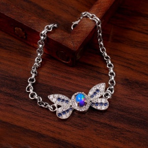 Puede incluir: Una pulsera de plata con un dije de mariposa con una piedra de ópalo azul e iridiscente rodeada de circonitas cúbicas transparentes y azules.
