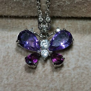 May include: A silver butterfly pendant necklace with purple and clear gemstones. The butterfly has two large purple pear-shaped stones and two smaller purple pear-shaped stones. The center of the butterfly has a cluster of clear round stones.