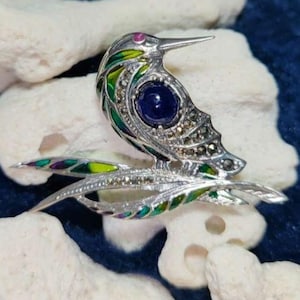 May include: A silver-toned hummingbird brooch with a blue gemstone eye and marcasite detailing. The bird is perched on a branch with green, yellow, and purple enamel leaves. A small pink gemstone accents the head.