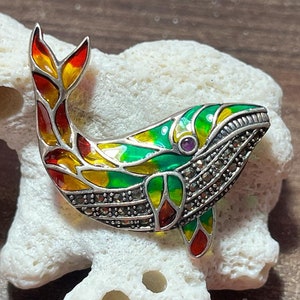 May include: A silver whale brooch with green, yellow, and red enamel and a purple gemstone eye. The whale is decorated with marcasite stones.
