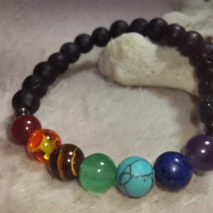 Puede incluir: Un brazalete de roca de lava negra con siete cuentas de piedras preciosas de colores. Las cuentas son rojas, naranjas, amarillas, verdes, azules, turquesas y moradas.