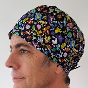 Op de afbeelding: Een zwarte scrub-muts met een kleurrijke print van verschillende Pokémon-personages. De muts heeft een knoop bovenop en een strik aan de achterkant.