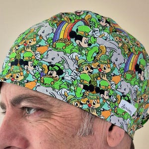 Peut inclure: Un bonnet de travail vert, blanc et noir avec un motif répété de Mickey Mouse et d'autres personnages de dessins animés portant des chapeaux verts et tenant des trèfles. Le bonnet a une étiquette blanche avec le texte "Scribblesy" dessus.