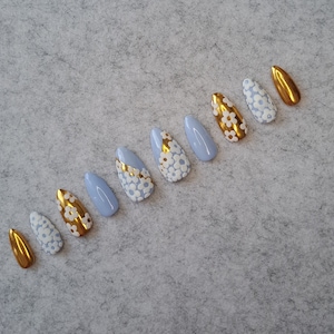 May include: A set of ten blue and gold press-on nails with a white floral design. The nails are in various sizes and shapes.