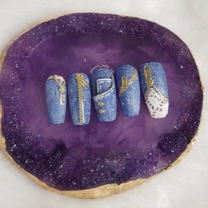 Puede incluir: Cinco u&ntilde;as postizas largas y ovaladas con un dise&ntilde;o de mezclilla. Cada u&ntilde;a tiene un detalle de cremallera dorada. Las u&ntilde;as se muestran en una piedra de &aacute;gata p&uacute;rpura y dorada.