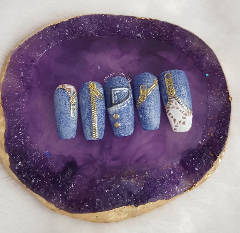 Puede incluir: Cinco u&ntilde;as postizas largas y ovaladas con un dise&ntilde;o de mezclilla. Cada u&ntilde;a tiene un detalle de cremallera dorada. Las u&ntilde;as se muestran en una piedra de &aacute;gata p&uacute;rpura y dorada.
