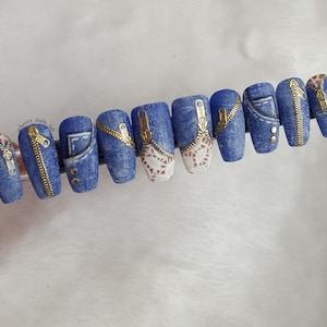Könnte beinhalten: Zehn blaue und weiße Kunstnägel mit goldenen Reißverschluss-Akzenten. Die Nägel sind mit einem Jeansmuster verziert und einige haben goldene Nieten.