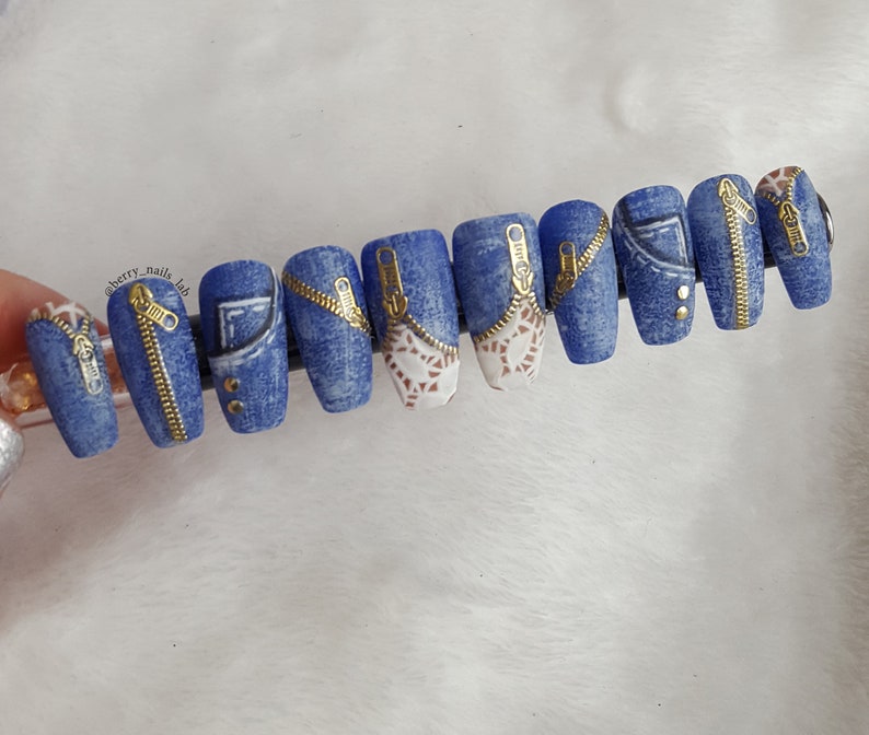 Puede incluir: Diez puntas de u&ntilde;as postizas azules y blancas con detalles de cremallera dorada. Las u&ntilde;as est&aacute;n decoradas con un patr&oacute;n de mezclilla y algunas tienen tachuelas doradas.