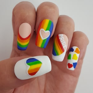 Pode incluir: Arte de unhas de orgulho arco-íris com pontas brancas e corações. As unhas apresentam um design de arco-íris com um coração branco em uma unha, a palavra "PRIDE" em outra e um coração de arco-íris em uma terceira unha. As unhas restantes têm uma base branca com corações de arco-íris.