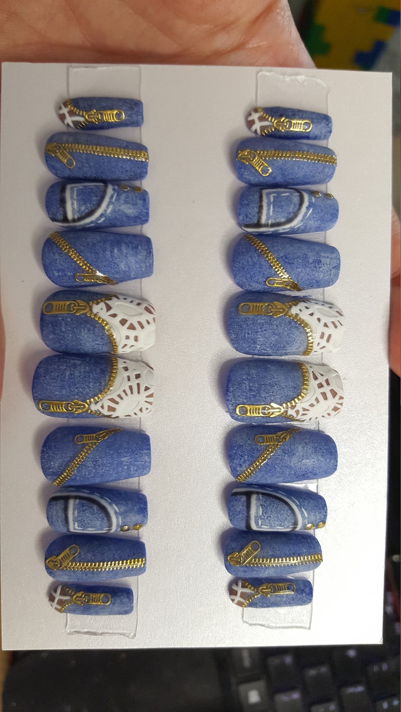 Puede incluir: Conjunto de diez u&ntilde;as postizas azules y blancas con detalles de cremallera dorada. Las u&ntilde;as est&aacute;n decoradas con un patr&oacute;n de mezclilla y encaje blanco.