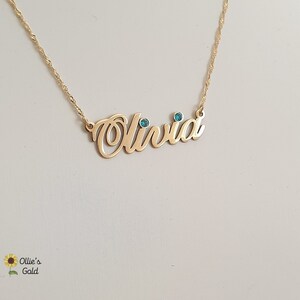 May include: Gold necklace with the name "Olivia" spelled out in cursive with two small blue gemstones.