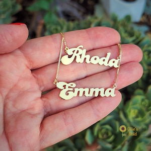 May include: Two gold necklaces with the names "Rhoda" and "Emma" in a cursive font. The necklaces are on a hand with a green background.