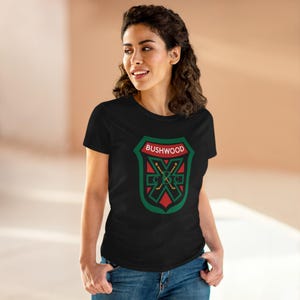 May include: Black t-shirt featuring a shield design with the text "BUSHWOOD" in red and white. The shield has a green border and a red and green emblem with crossed hockey sticks. The shirt is made of a soft material.