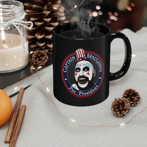 Puede incluir: Taza de café negra con un gráfico circular blanco que presenta un payaso con barba y un sombrero rojo, blanco y azul. El texto "Captain Spaulding For President" está escrito alrededor del círculo.