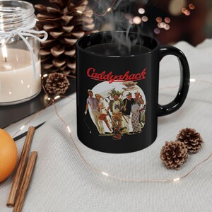 May include: Black ceramic mug featuring the word "Caddyshack" in red script above a graphic of several people. The mug has a curved handle and is filled with a dark liquid, possibly coffee or tea.