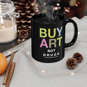 Peut inclure: Une tasse à café noire avec du texte blanc qui dit "BUY ART NOT DRUGS" en lettres colorées. La tasse est sur une surface gris clair avec des pommes de pin et des lumières de fée.