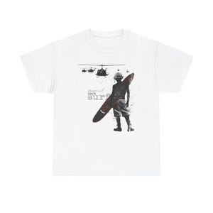 May include: White t-shirt featuring a graphic of a soldier holding a surfboard, with helicopters in the background. The text "Charlie don't surf" is printed on the shirt in a distressed font. The design is in shades of gray and red.