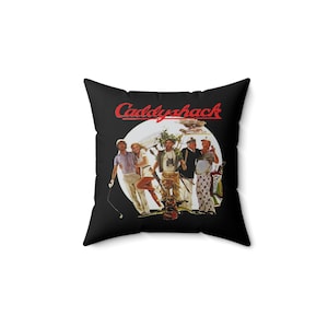 May include: A square black throw pillow featuring the red text Caddyshack above a circular graphic illustration of five figures in golf attire, including a person holding a golf club, a person in a white outfit, and a person with binoculars.