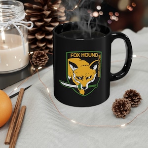 Puede incluir: Taza de café negra con un logotipo verde, amarillo y blanco que dice "FOX HOUND SPECIAL FORCE GROUP" y un zorro de dibujos animados con un cuchillo en la boca.